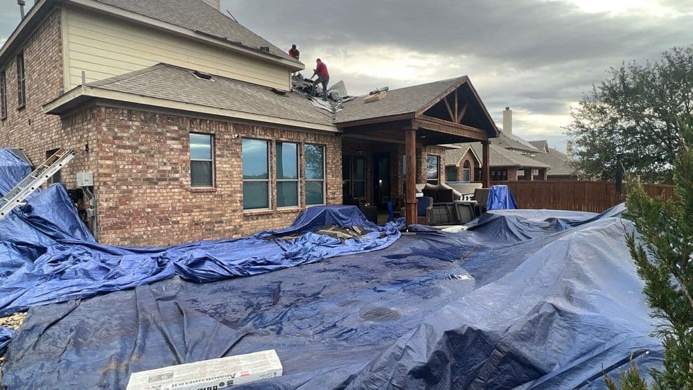 Roofing workers installing shingles on a home, covered with tarps in a cloudy sky.