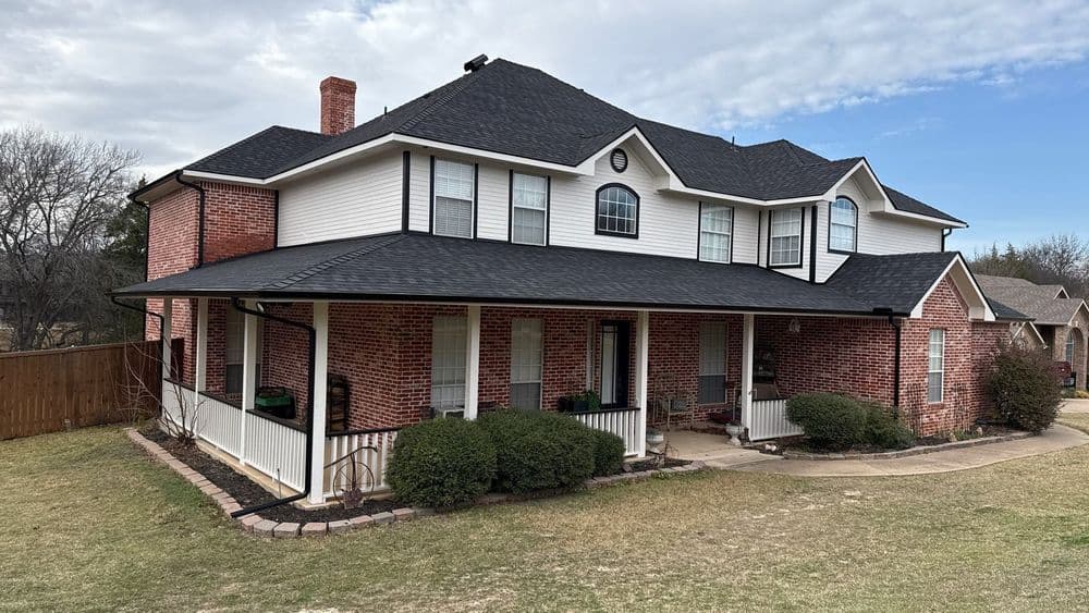 Two-story brick home with a large porch and landscaped yard under a cloudy sky.
